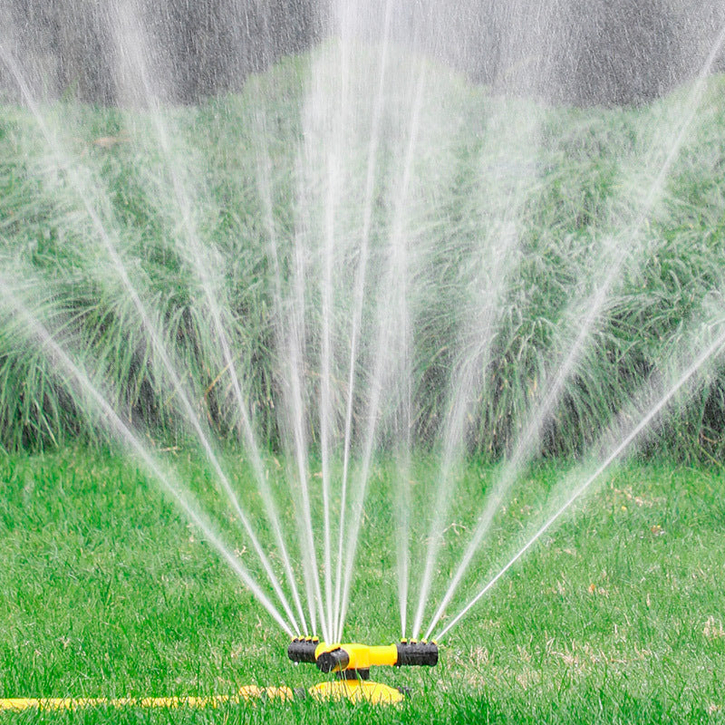 Yellow 360-degree swivel lawn sprinkler spraying water in a fan pattern over green grass and landscaping.