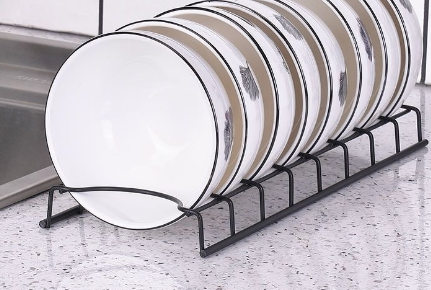 Black wire dish rack holding a row of white and tan bowls with black rims on a speckled kitchen countertop.