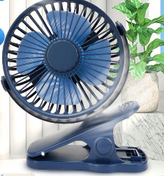 Dark blue portable clip-on fan with three blades, resting on a white surface next to a potted green plant.