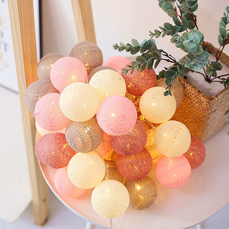 String of glowing cotton ball lanterns in shades of blush, cream, and taupe arranged in a pile on a small white table.