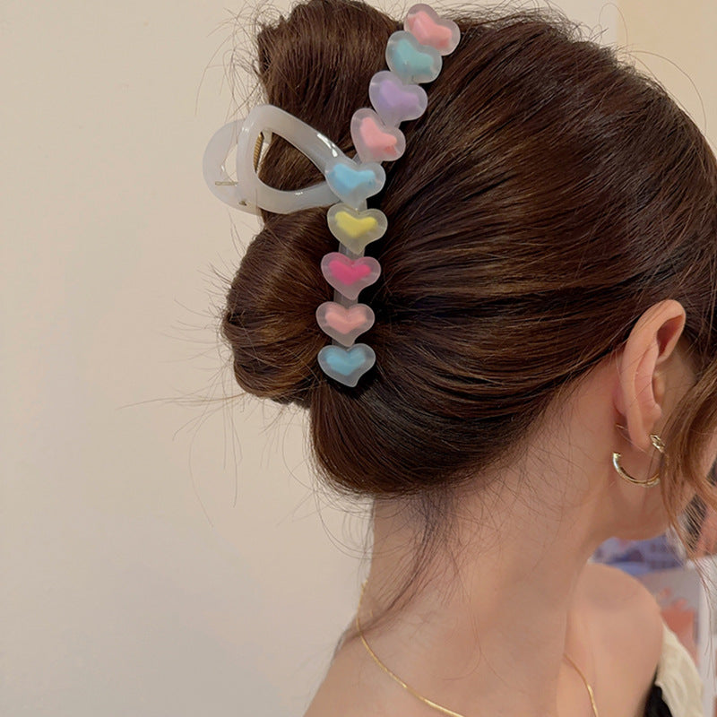 Clear claw hair clip featuring a row of translucent, pastel-colored hearts, holding a brown hair bun in place.