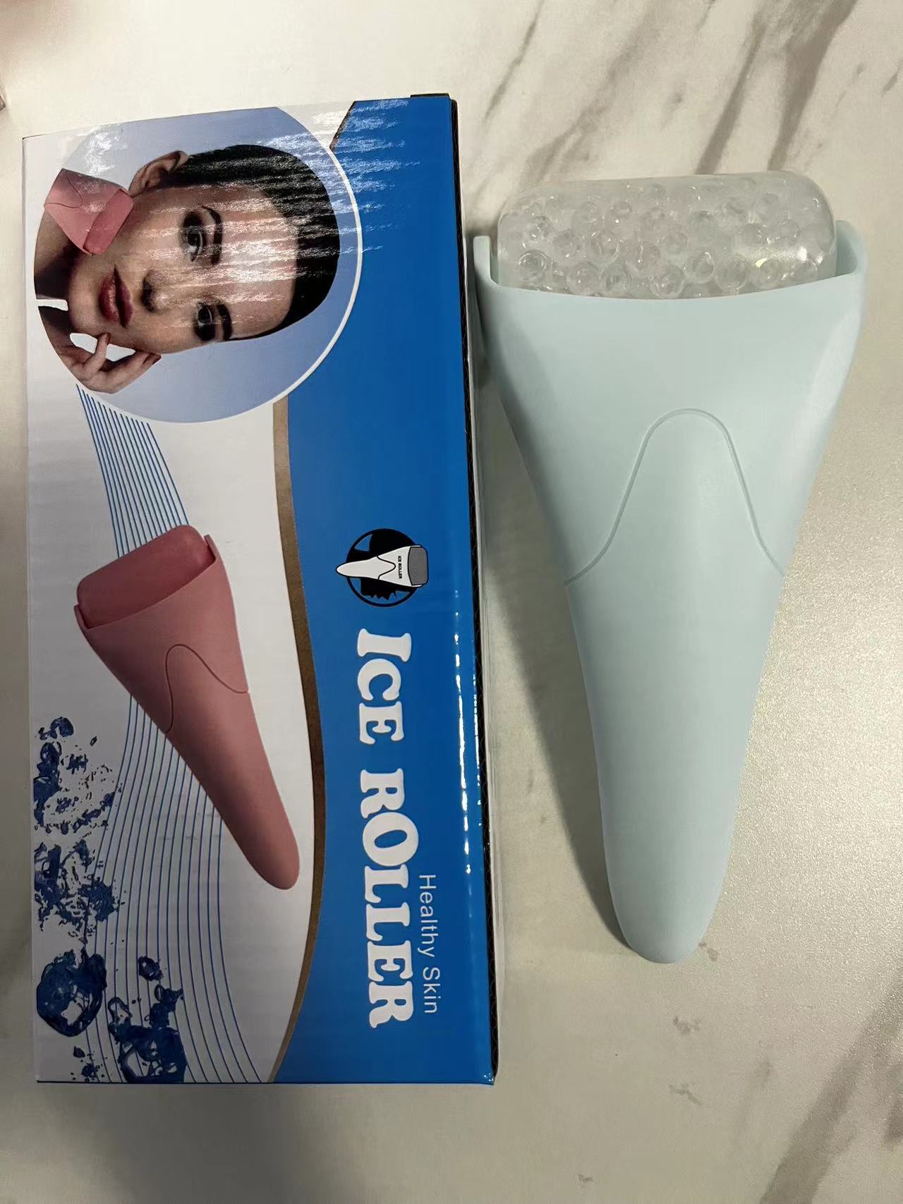 Light blue facial ice roller with a textured, studded drum, shown next to its retail box featuring a pink roller.