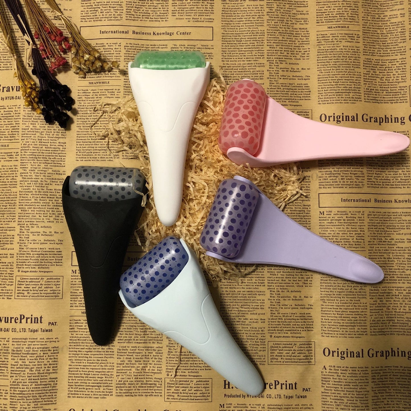 Five Chill Glide facial ice rollers in white, pink, black, purple, and blue arranged on vintage newspaper.