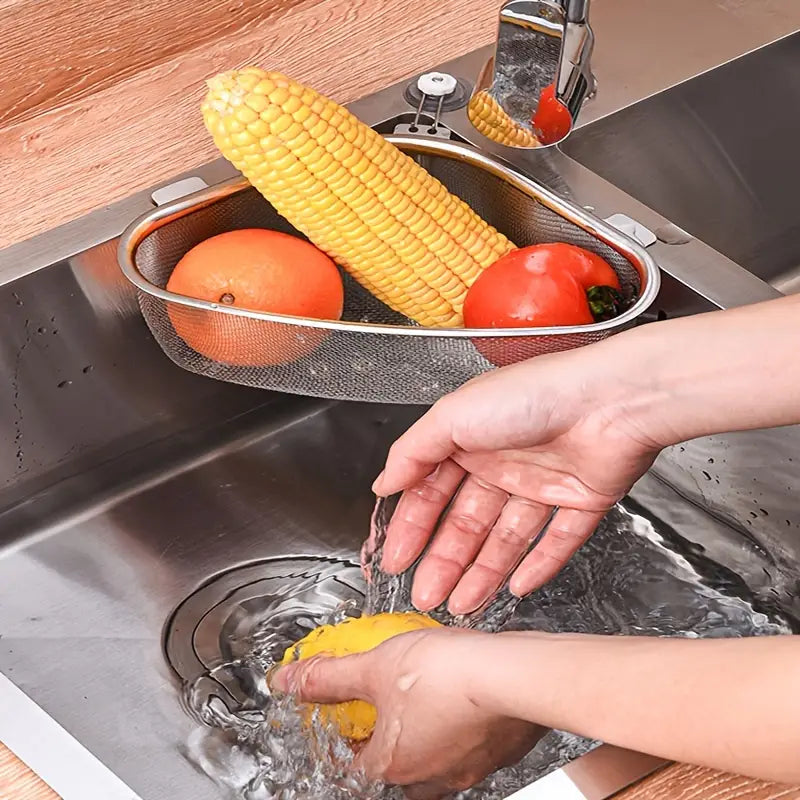 Triangle stainless steel mesh sink strainer holding corn, an orange, and a tomato, mounted in a steel kitchen sink.