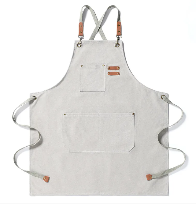 Light grey canvas apron with cross-back straps, tan leather accents, brass hardware, and multiple pockets.