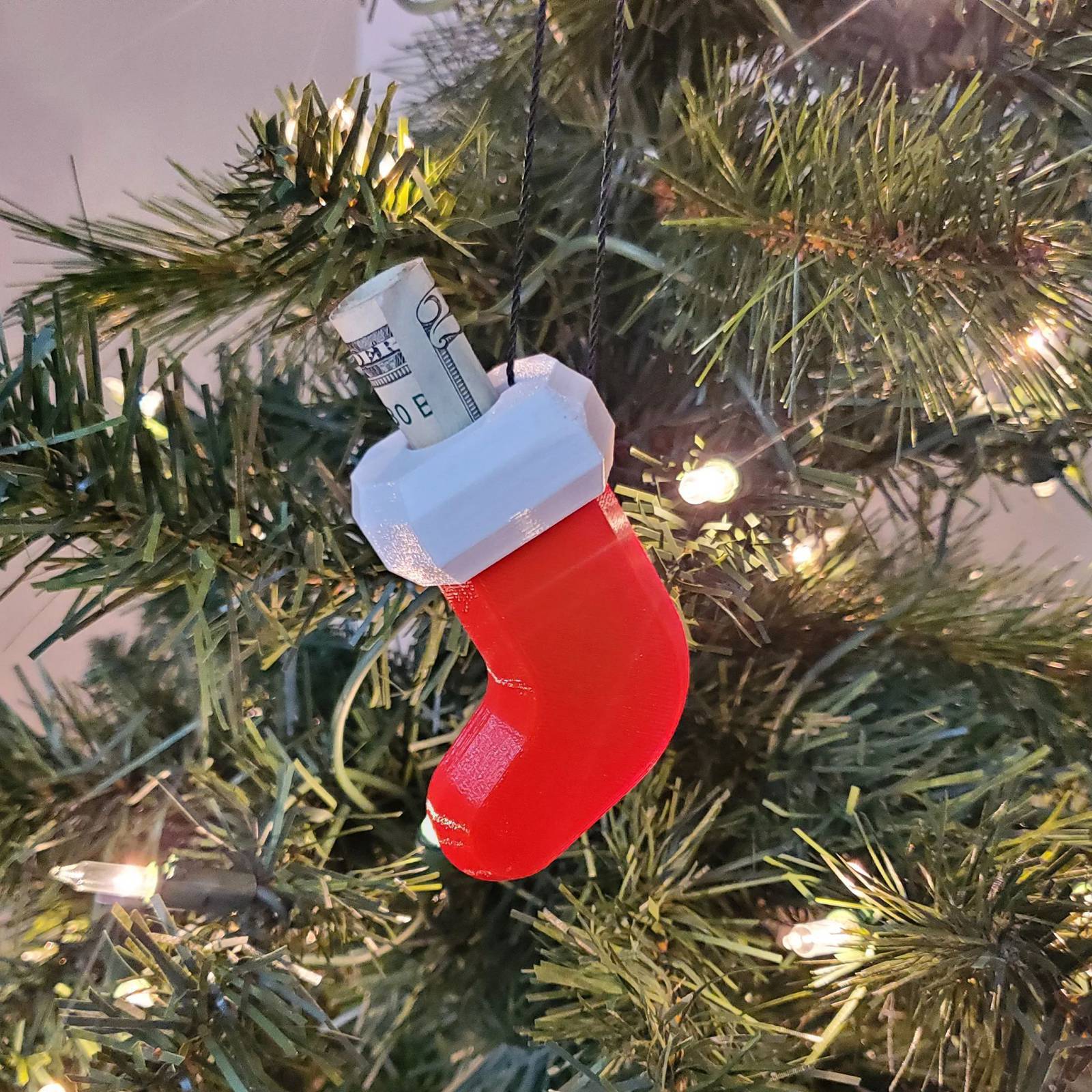 Red and white 3D-printed stocking-shaped ornament hanging on a Christmas tree with a rolled-up dollar bill inside.