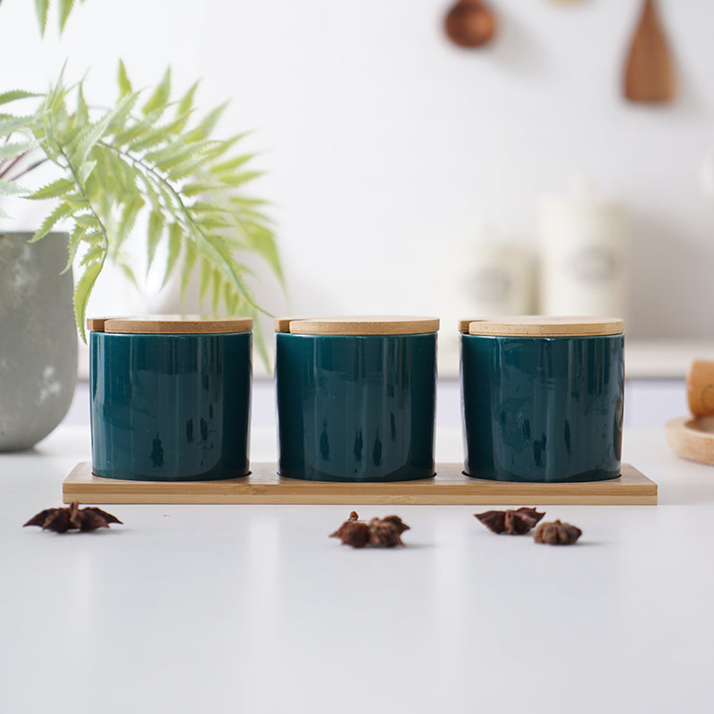 Three glossy dark teal ceramic spice jars with bamboo lids on a matching wooden tray, set on a white kitchen counter.