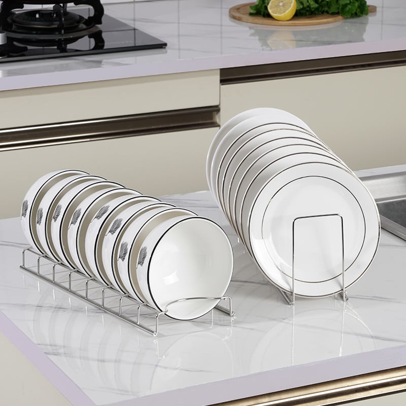 Sleek stainless steel organizers holding stacks of white bowls and plates with delicate feather patterns on marble.