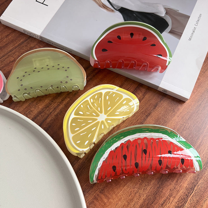 Four semi-circular fruit-themed acrylic hair claws shaped like watermelon, kiwi, and lemon slices on a wooden surface.