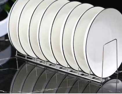 Sleek stainless steel dish rack holding a row of white plates with thin black rims on a black marble surface.
