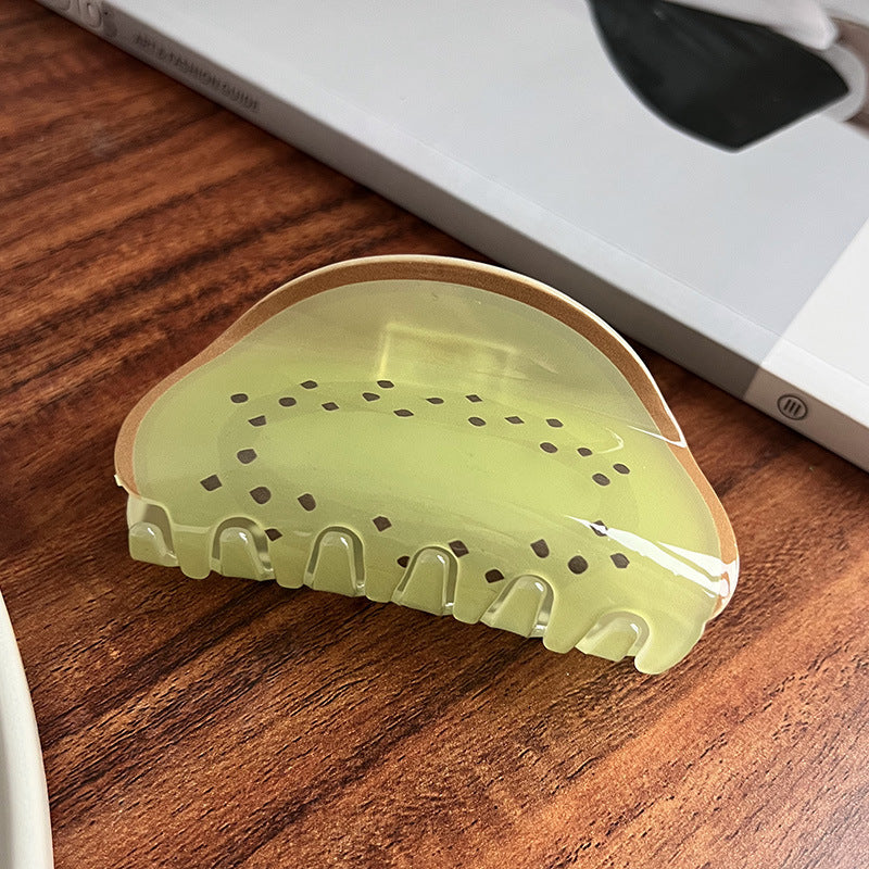 Translucent, light green acrylic hair claw shaped like a kiwi slice with brown seeds, resting on a wooden surface.