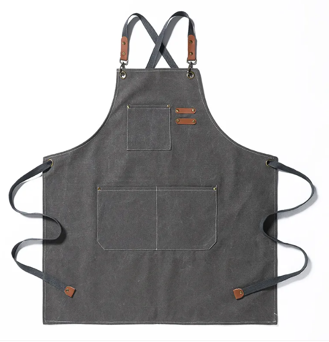 Charcoal grey canvas apron with brown leather accents, cross-back straps, and multiple utility pockets.