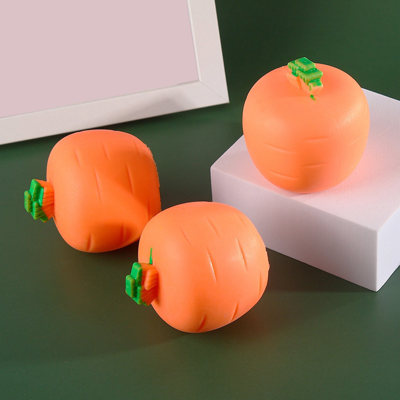Three round, orange carrot-shaped stress relief toys with small green tops, arranged on white blocks against green.
