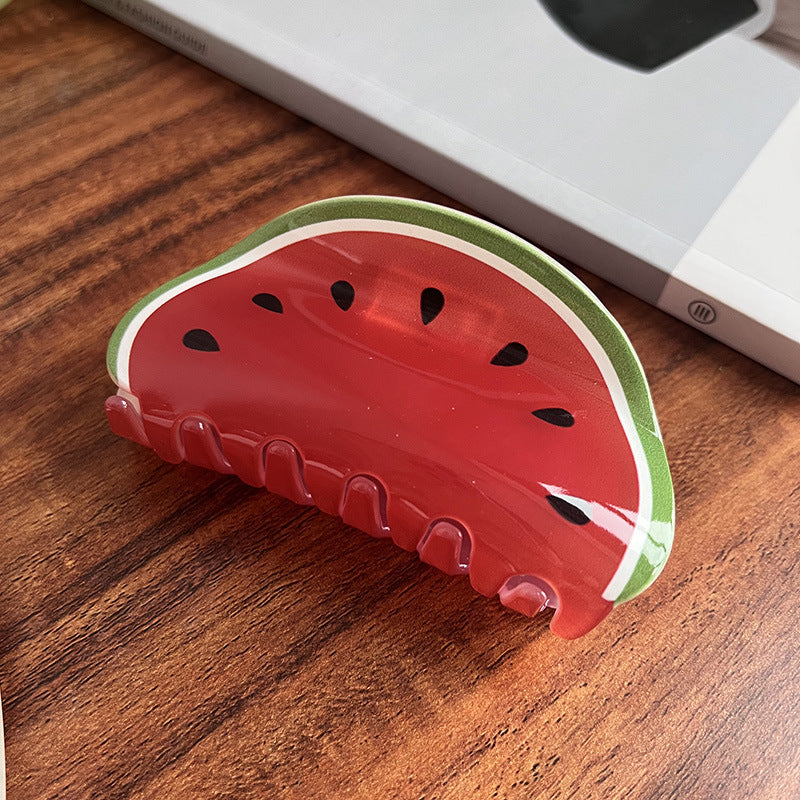 Watermelon-shaped acrylic hair claw clip with red fruit pattern, black seeds, and a green rind on a wooden table.