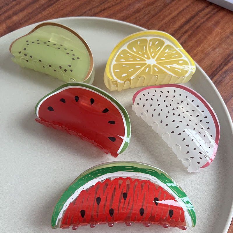 Five fruit-themed acrylic hair claws shaped like kiwi, lemon, watermelon, and dragon fruit on a white plate.