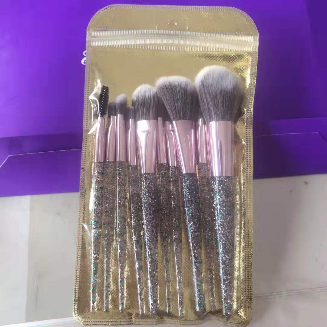 Ten-piece makeup brush set with glitter handles, rose gold ferrules, and soft white-tipped bristles in a gold pouch.