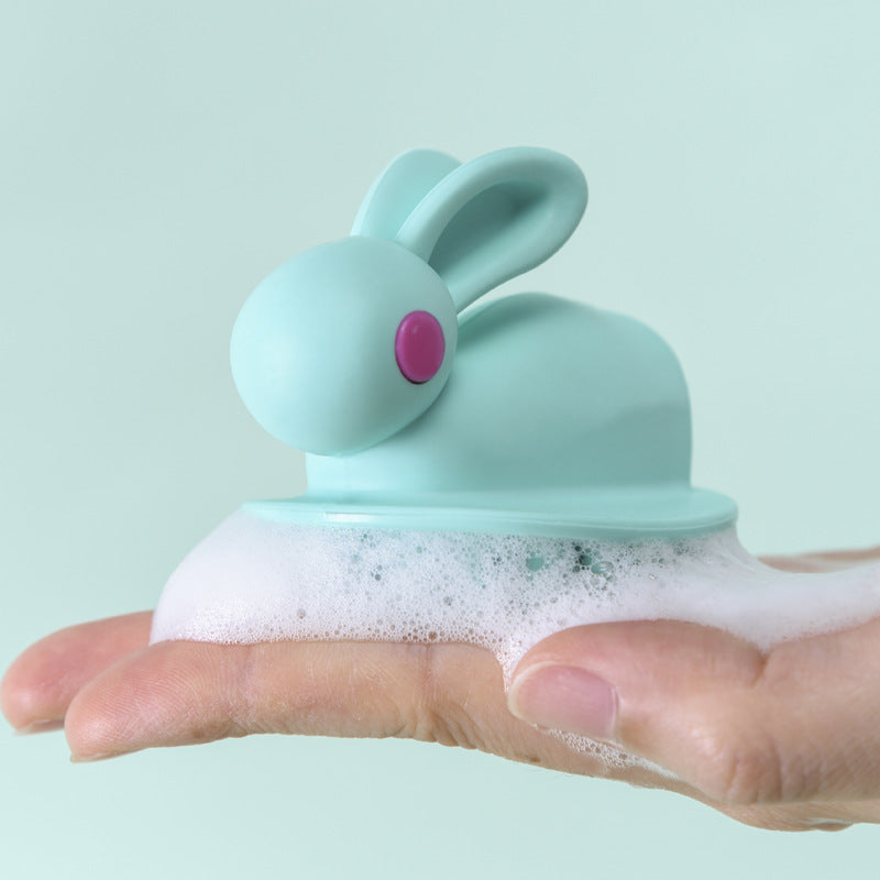 Light blue, rabbit-shaped kids' scalp brush with a pink eye, shown lathered with soap on a hand.