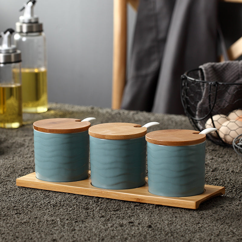 Three blue Nordic-style ceramic spice jars with wooden lids and spoons on a matching rectangular bamboo tray.