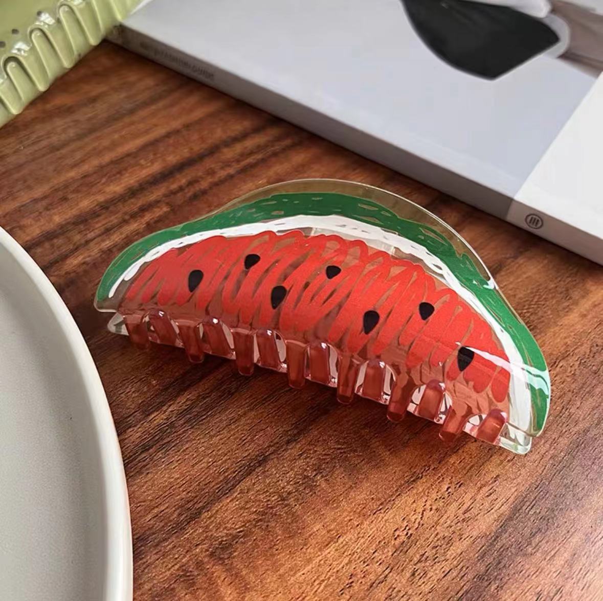 Watermelon-shaped acrylic hair claw with red, white, and green details and black seed accents.