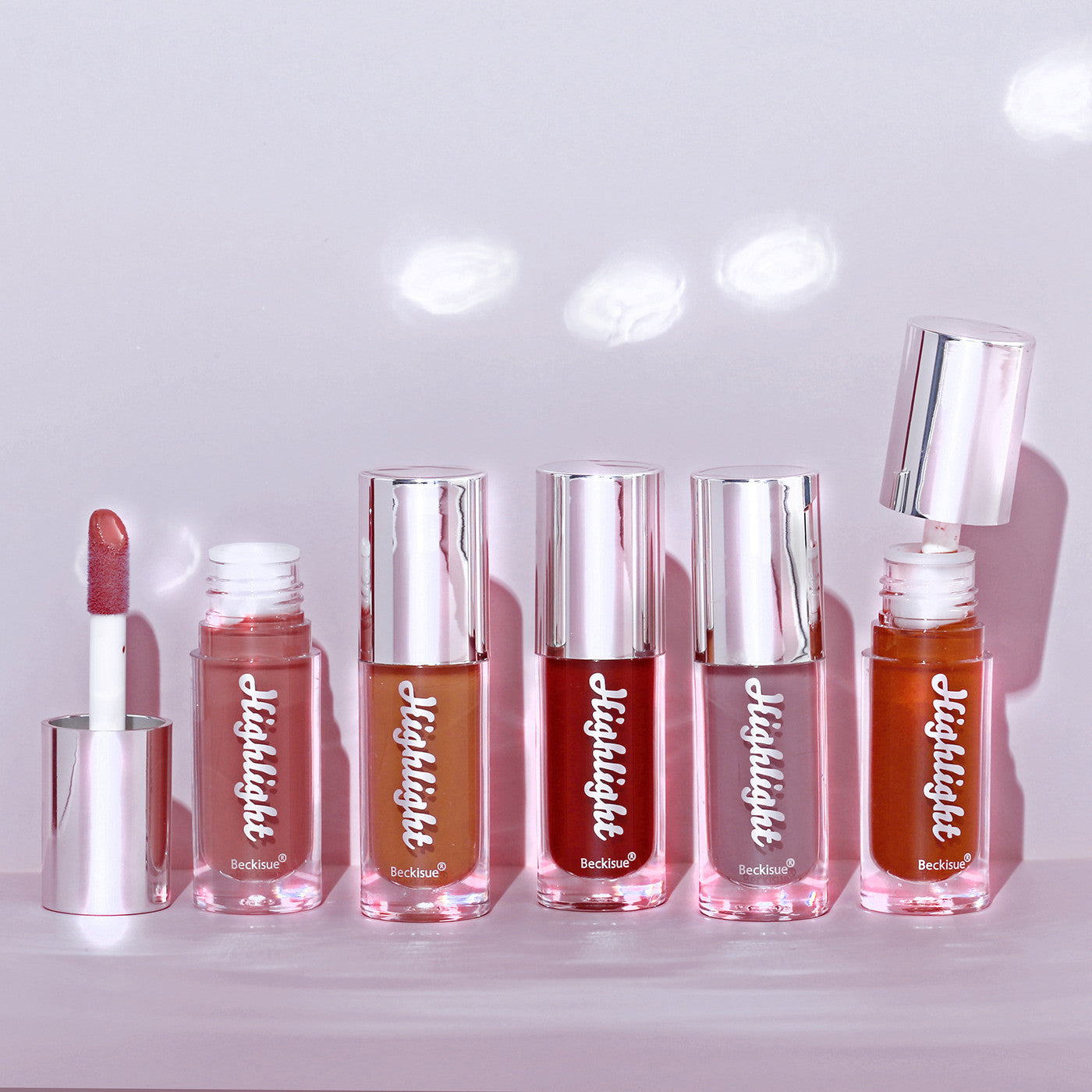 Six Beckisue Highlight lip gloss tubes in various shades with shiny silver caps on a soft, light purple background.