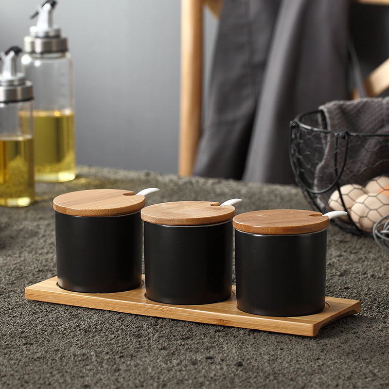 Three matte black ceramic spice jars with bamboo lids and spoons, organized on a matching bamboo serving tray.