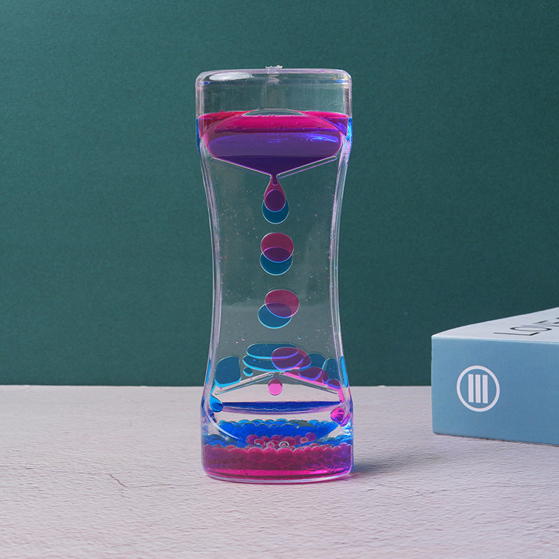 Clear acrylic liquid motion timer with floating pink and blue droplets on a textured white surface.