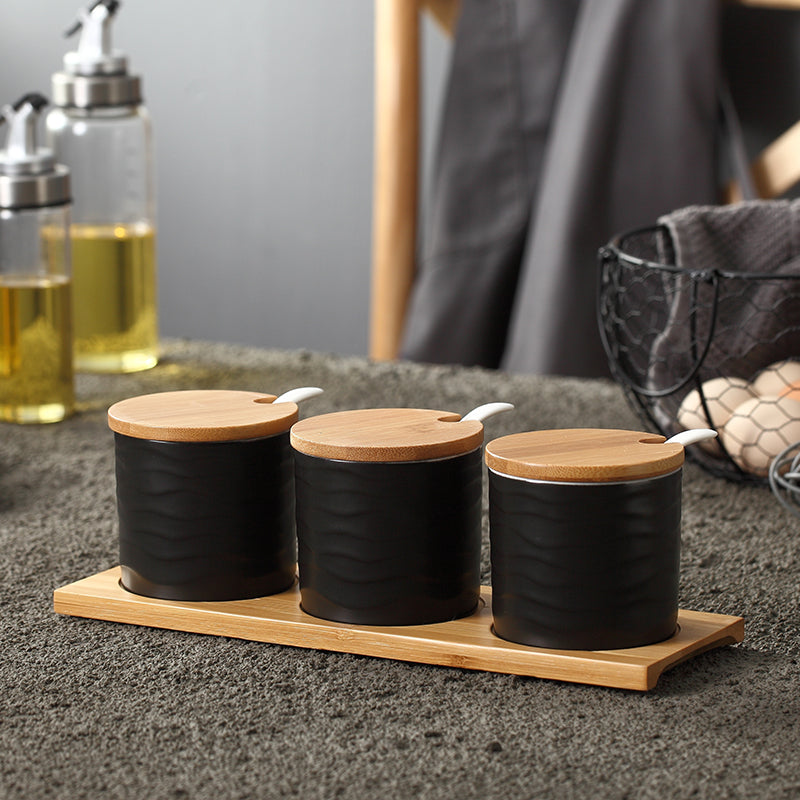 Three black ribbed ceramic spice jars with bamboo lids and spoons on a matching wooden tray, Nordic minimalist style.