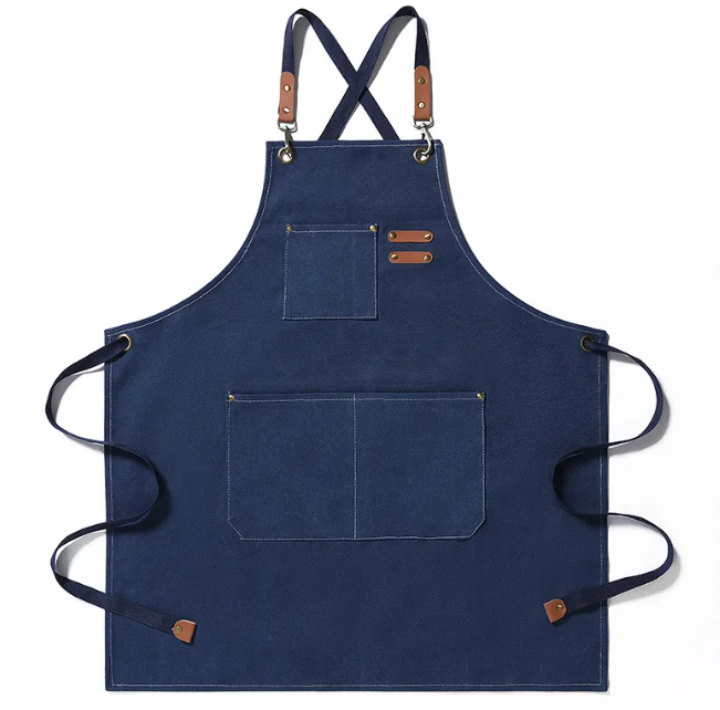 Navy blue canvas apron with cross-back straps, tan leather accents, brass hardware, and multiple pockets.