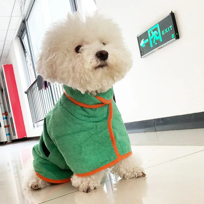 Fluffy white dog wearing a green quick-dry bathrobe with orange trim, sitting indoors on a tiled floor.